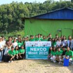 A group of people posing for a photo outdoors in front of a green and blue building with a banner that reads "NExCO Meet 2025."