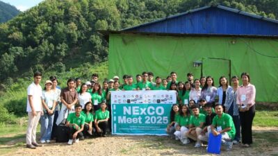 A group of people posing for a photo outdoors in front of a green and blue building with a banner that reads "NExCO Meet 2025."