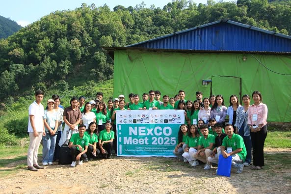 A group of people posing for a photo outdoors in front of a green and blue building with a banner that reads "NExCO Meet 2025."