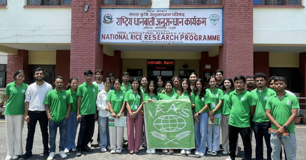 A wide shot of the IAAS Nepal National Congress opening ceremony with a panel of speakers on an outdoor stage addressing a large, applauding audience of agricultural students. The scene is set on a university campus with farm fields and green mountains in the background.