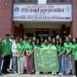 A wide shot of the IAAS Nepal National Congress opening ceremony with a panel of speakers on an outdoor stage addressing a large, applauding audience of agricultural students. The scene is set on a university campus with farm fields and green mountains in the background.
