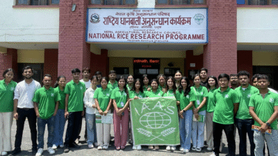 A wide shot of the IAAS Nepal National Congress opening ceremony with a panel of speakers on an outdoor stage addressing a large, applauding audience of agricultural students. The scene is set on a university campus with farm fields and green mountains in the background.